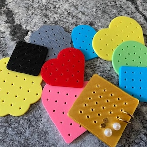 Op de afbeelding: Een verzameling veelkleurige plastic pegboard-organizers in diverse vormen, waaronder harten, vierkanten, cirkels en geschulpte randen. Elk stuk heeft een raster van kleine gaatjes, met één goudkleurig vierkant waar twee gouden oorringen in zitten.