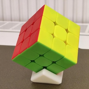 May include: A multi-coloured 3x3 puzzle cube featuring red, yellow, and green faces, balanced on a small white triangular plastic stand. The cube has rounded edges on each individual square segment and is positioned at an angle to display three sides simultaneously.