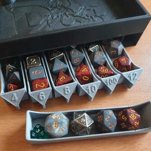 May include: A black 3D-printed storage box with multiple light grey compartments labelled with numbers 4, 6, 8, 10, 100, and 12. The compartments contain various multi-sided polyhedral dice in colours including red, grey, silver, and green with gold numbering.