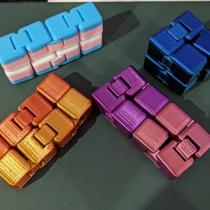 Pode incluir: Quatro brinquedos fidget tipo cubo infinito retangulares feitos de plástico. Um cubo apresenta listras horizontais em azul claro, rosa e branco. Os outros três cubos são de cores metálicas sólidas: um azul profundo, um laranja dourado e um roxo magenta.
