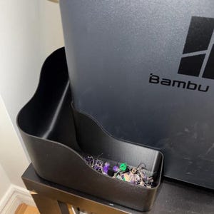 May include: A black plastic storage bin with a curved side profile attached to the side of a dark gray Bambu 3D printer. The bin contains a small collection of tangled purple, green, and black plastic filament waste scraps from the printing process.