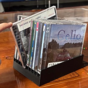 Può includere: Un supporto da scrivania in plastica nera per CD contenente diverse custodie per dischi compatti. La custodia in primo piano presenta una copertina con il testo Cello the most relaxing album in the world... ever! e un'auto rossa.