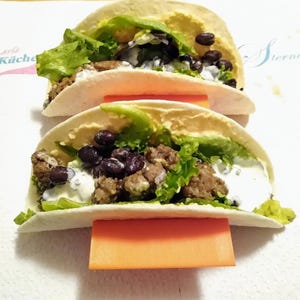 May include: Two soft flour tortillas filled with seasoned ground meat, black beans, fresh green lettuce, and a white creamy sauce. Each taco is held upright by a bright orange plastic stand, resting on a white surface with partial text.