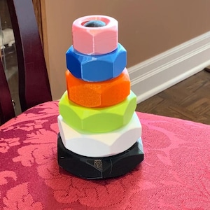 May include: A stack of six hexagonal nuts in descending order of size, featuring a color gradient from top to bottom in light pink, blue, orange, lime green, white, and black. The nuts are arranged vertically to form a tower-like structure.