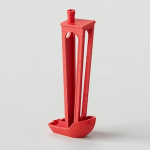 Pode incluir: Um objeto vermelho brilhante impresso em 3D com uma base em forma de pequeno barco ligada a uma torre vertical de quatro lados, com recortes em arco em cada lado e uma pequena saliência cilíndrica na plataforma quadrada superior.