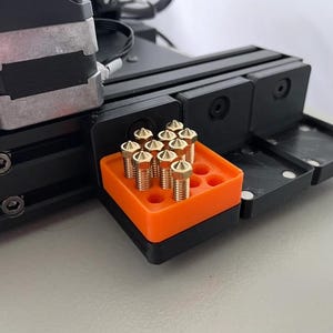 May include: A 3D printer nozzle storage organizer featuring a bright orange rectangular base with multiple circular slots. Several metallic gold-colored brass nozzles are inserted into the slots. The organizer is attached to the black metal frame of a 3D printer.