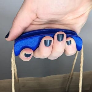May include: A blue plastic bag handle grip with a textured surface and a curved shape. The grip features two slots on the underside for the paper bag's twisted brown paper handles to pass through, providing a comfortable hold for carrying.