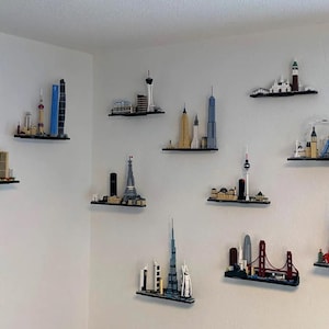 May include: A collection of twelve individual LEGO Architecture skyline sets mounted on small black wall shelves. The sets feature various global city landmarks, including the Eiffel Tower, Burj Khalifa, Golden Gate Bridge, and Tower Bridge.