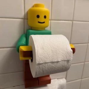 Op de afbeelding: Een aan de muur gemonteerde toiletrolhouder in de vorm van een geel, groen en bruin plastic speelgoedfiguur. Het figuur heeft een cilindervormig geel hoofd met een simpel zwart smiley-gezicht en houdt een rol wit toiletpapier met reliëf vast.