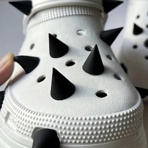 May include: A white clog shoe featuring a series of matte black conical spikes attached to the upper surface. The shoe has a textured exterior with circular ventilation holes and a pivoting heel strap, also adorned with black spikes.