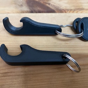 Könnte beinhalten: Zwei schwarze, 3D-gedruckte Flaschenöffner in Form von Schlüsselanhängern. Jeder Öffner verfügt über ein gebogenes Hakendesign zum Entfernen von Kronkorken und ein kreisförmiges Loch an einem Ende zur Befestigung an einem Metallring. Einer ist an Schlüsseln befestigt.