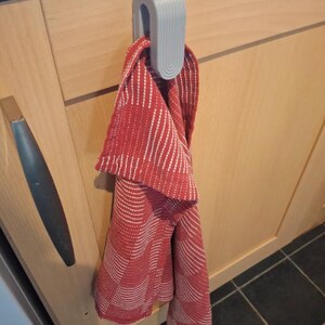 May include: A light gray U-shaped plastic cabinet hook hangs over the top edge of a wooden kitchen cabinet door. A red and white patterned kitchen towel is draped through the hook, hanging vertically down the front of the cabinet door.