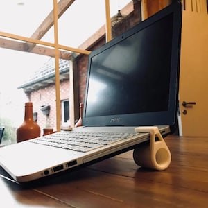 May include: A silver and black laptop computer is elevated by a white, cylindrical, 3D-printed plastic stand. The stand features a circular opening and a hook-like top that secures the edge of the laptop, creating an angled typing position.