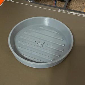 May include: A circular grey tray with a raised rim and a series of parallel horizontal ridges along the interior base. In the center of the tray, there is a small, embossed logo featuring two curved lines that resemble a stylized letter K.