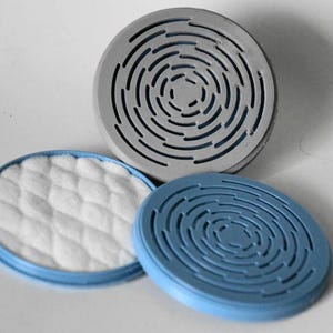 May include: Three circular components including one light gray disc with a concentric slotted pattern, one light blue disc with a matching slotted pattern, and one light blue circular base containing a white, quilted, textured fabric insert.