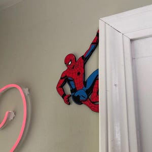 May include: A flat, wall-mounted cutout of the comic book character Spider-Man, depicted in a classic red and blue suit with black web detailing. The figure is shown in a dynamic crouching pose, with one arm extended upwards as if climbing.
