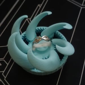 May include: A silver-toned infinity knot ring resting on a light blue, 3D-printed circular display stand. The stand features a series of curved, tentacle-like protrusions radiating outward from the center, providing a secure base for the ring.