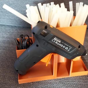 Könnte beinhalten: Eine schwarze kabellose Heißklebepistole mit eingesetztem weißem Klebestift, die auf einem orangefarbenen Kunststoff-Organizer steht. Der Ständer enthält weitere weiße Klebestifte und ein schwarzes Ladekabel. Die Klebepistole trägt den Text Made in China.