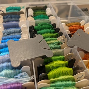 May include: A clear plastic storage container filled with embroidery floss wound onto white and gray plastic bobbins. The bobbins are organized in rows by color, featuring shades of blue, green, yellow, orange, and red, with some bobbins labeled.