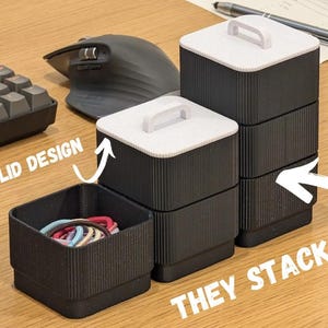 Puede incluir: Un conjunto de recipientes de almacenamiento cuadrados negros con lados acanalados y tapas blancas con pequeñas asas. Los recipientes son modulares y apilables. Un recipiente está abierto y contiene gomas para el pelo. Texto: NEW LID DESIGN y THEY STACK.