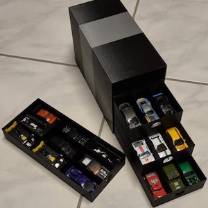 May include: A black storage tower with a gray horizontal stripe featuring three pull-out drawers, each containing multiple miniature toy cars in various colors including red, yellow, green, blue, and white, alongside a separate matching black tray.