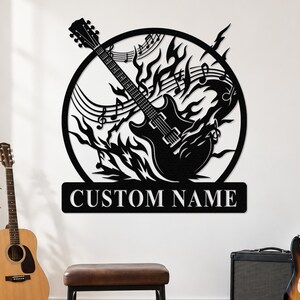 May include: A black metal wall art piece featuring a circular frame with a guitar silhouette surrounded by flames and musical notes. Below the circle is a rectangular plaque with the text CUSTOM NAME in bold, capitalised, white-outlined letters.