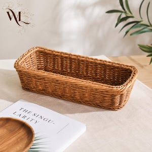 May include: A rectangular woven basket with a light brown finish and a structured rim. The basket features a consistent, tight weave pattern throughout its sides and base, creating a sturdy, open-top container for storage or display purposes.