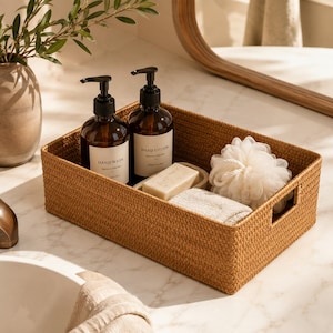 May include: A rectangular woven storage basket in a warm tan color containing two amber glass pump bottles labeled Hand Wash and Hand Lotion, a rectangular bar of soap, a folded beige washcloth, and a white mesh bath sponge.