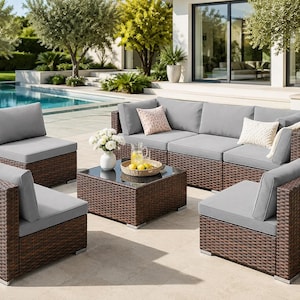 Peut inclure: Un ensemble de mobilier de jardin en osier marron comprenant un canapé modulaire, deux fauteuils et une table basse carrée avec un plateau en verre. L'ensemble inclut des coussins gris clair, deux coussins décoratifs et un plaid beige.
