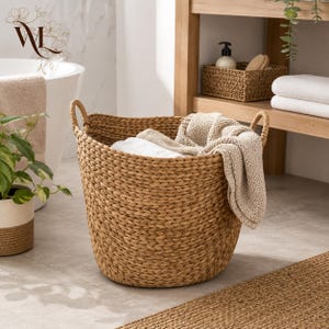May include: A round, woven seagrass basket with two integrated handles, containing a folded beige knit blanket. The basket features a textured, braided pattern and is positioned in a bathroom setting with a white tub and wooden shelving.