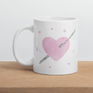 Op de afbeelding: Een witte keramische mok met een groot, zachtroze hart in het midden. Een zwart-witte lijntekening van een dwarsfluit is diagonaal over het hart geplaatst. Kleine roze hartjes en zwarte muzieknoten zijn verspreid rondom het ontwerp.