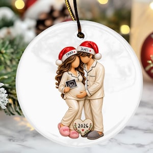 May include: A round, clear acrylic Christmas ornament featuring a couple in pajamas and Santa hats. The woman holds an ultrasound image, and the man stands beside her. The hats are personalized with the names Emma and James. A heart-shaped plaque at their feet displays the year 2026.