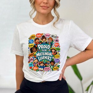 May include: A white short-sleeved t-shirt featuring a colorful graphic print with the text Proud To Be Multilingual Learners. The design includes illustrations of diverse children, a globe, and various international greetings and flags.