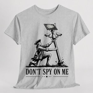May include: A light gray t-shirt featuring a black graphic of a person in 18th-century attire kneeling and holding a tall pole topped with a solar panel and two security cameras. The text DON'T SPY ON ME is printed in black below the graphic.