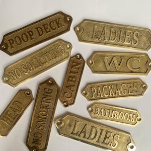 Pode incluir: Uma coleção de nove placas retangulares de latão com cantos chanfrados e orifícios de montagem. As placas apresentam letras em relevo com o texto: POOP DECK, LADIES, NO SOLICITING, WC, HEAD, NO SMOKING, CABIN, PACKAGES, BATHROOM e LADIES.