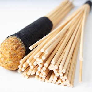 Puede incluir: Primer plano de un instrumento de percusión de madera con un mango negro y un haz de palos de madera. Los palos están agrupados y tienen un color madera natural.