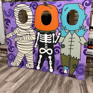 May include: A large rectangular Halloween photo prop board featuring a purple background with swirling patterns. It displays three painted characters: a white mummy, a skeleton with a pumpkin head, and a blue-skinned zombie, each with a cutout hole.