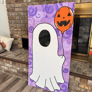 May include: A rectangular wooden board featuring a painted white ghost with a circular cutout for a face. The ghost holds an orange jack-o'-lantern balloon. The background is painted with purple swirls, creating a festive Halloween-themed display.