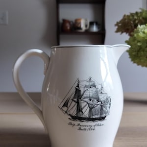 May include: A white ceramic pitcher featuring a black and white illustration of a sailing ship. The text below the ship reads Ship Recovery of Salem Built 1794. The pitcher has a curved handle and a pouring spout with a thin black rim detail.