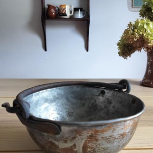 May include: A round metal bowl with a weathered, silver-toned interior and a copper-colored exterior finish. The bowl features a thick, dark, rusted iron swing handle attached to the sides with metal loops, showing signs of age and oxidation.