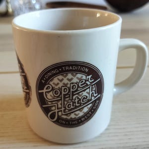Puede incluir: Una taza de cerámica blanca con un logotipo circular marrón en la parte frontal. El logotipo contiene el texto Copper Hatch en una fuente manuscrita estilizada, rodeado por las palabras A DINING TRADITION y ON THE WEST SIDE, con un patrón geométrico.