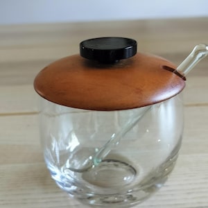May include: A clear glass condiment jar with a rounded body, featuring a circular wooden lid topped with a small black knob. A clear glass spoon is inserted through a notch in the wooden lid, resting inside the jar. The design is simple and functional.