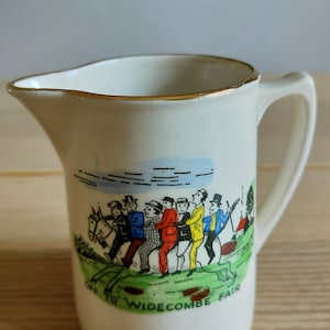 May include: A cream-colored ceramic creamer with a gold-trimmed rim and handle. The front features a colorful illustration of seven figures riding a horse, with the text "OFF TO WIDECOMBE FAIR" printed in green along the bottom of the graphic.