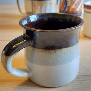May include: A ceramic mug featuring a tri-color glaze with a dark brown rim, a light gray middle section, and a white base. The mug has a curved handle and a smooth, glossy finish. Other mugs are visible in the blurred background behind it.