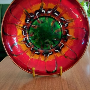 May include: A round metal bowl featuring a vibrant enameled interior with a deep green center, transitioning into bright orange and red bands. The surface displays dark, flowing, abstract streaks radiating outward toward the copper-toned rim.