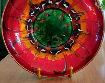 May include: A round metal bowl featuring a vibrant enameled interior with a deep green center, transitioning into bright orange and red bands. The surface displays dark, flowing, abstract streaks radiating outward toward the copper-toned rim.