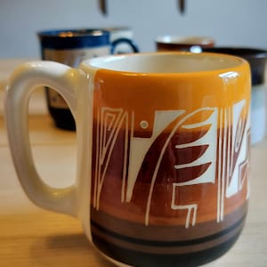 Può includere: Una tazza in ceramica con un design a blocchi di colore orizzontali nelle tonalità dell'arancione, marrone e nero. L'esterno è decorato con motivi geometrici e a foglia incisi in bianco. La tazza presenta un manico spesso e curvo.