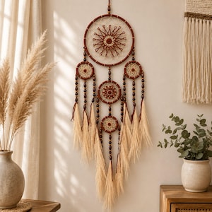 May include: A large wall-hanging dreamcatcher featuring a central brown hoop with a star-shaped woven web, accented by four smaller hoops below. The structure is decorated with dark brown wooden beads and long, light beige feathery fringe tassels.
