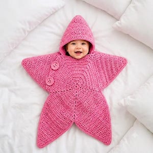 May include: A bright pink crocheted star-shaped bunting with a pointed hood and two large matching pink buttons on the front. The garment features a textured yarn construction and is designed to wrap around the body in a five-pointed star silhouette.
