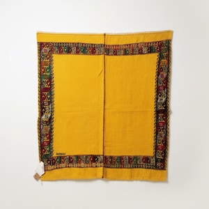 May include: A rectangular yellow textile featuring a wide border with a repeating geometric pattern in red, green, and dark brown. The fabric has a visible vertical seam running down the center and a small white tag attached to the bottom left corner.